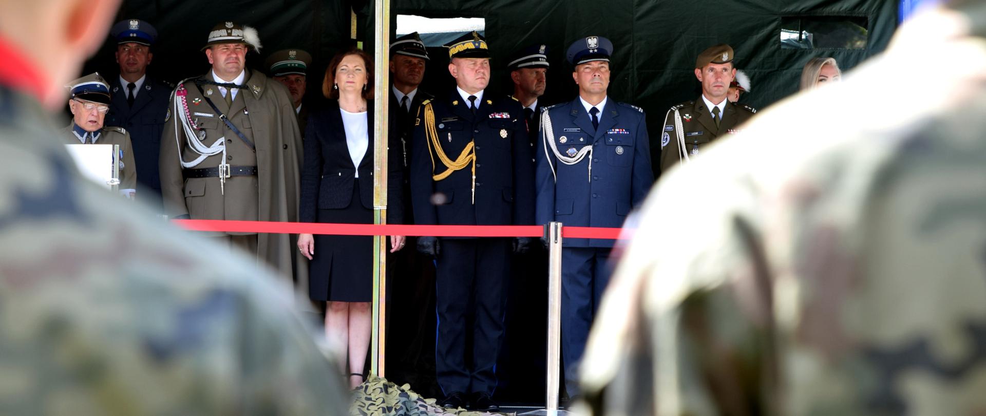 Zdjęcie zrobione na zewnątrz podczas uroczystego apelu. Pod zadaszeniem stoją zaproszeni honorowi goście. Wśród nich generał Państwowej Straży Pożarnej oraz oficerowie Wojska Polskiego, Policji, Obrony Terytorialnej Służby Więziennej. 