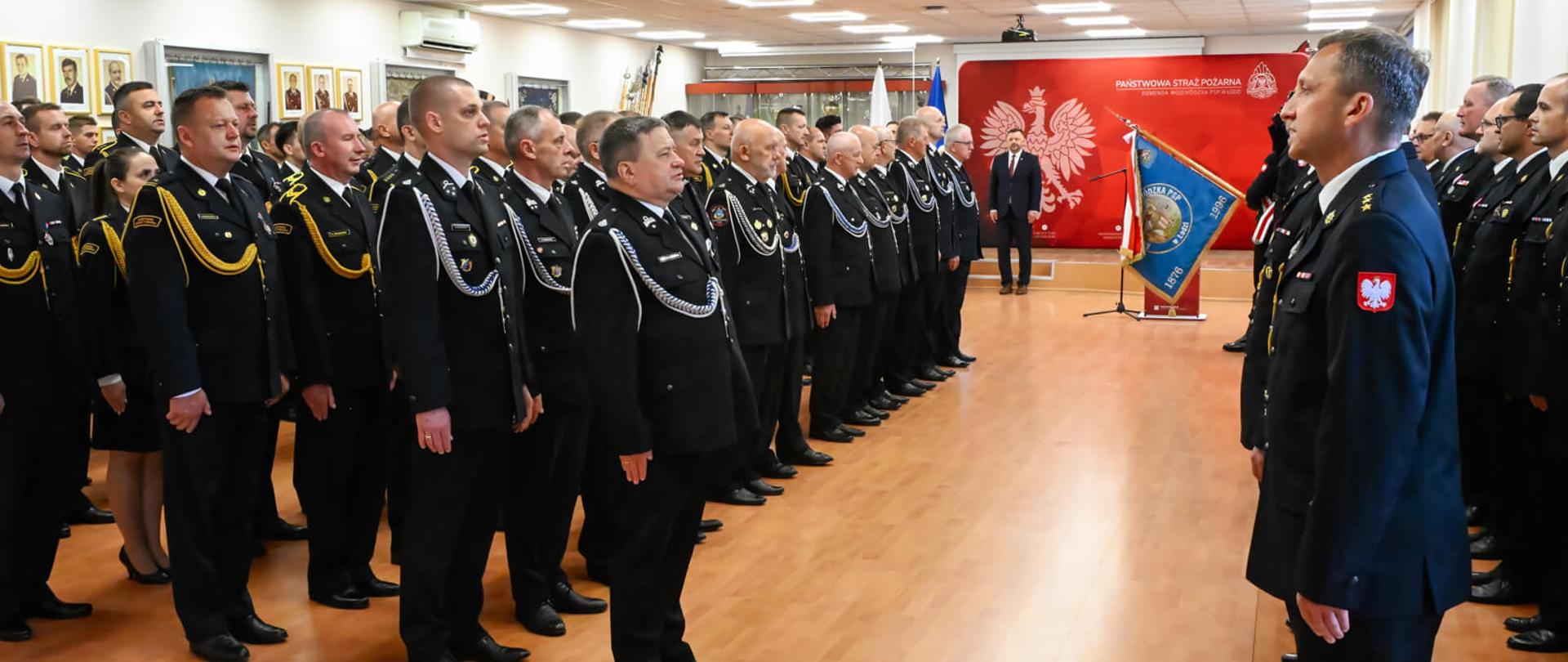 Uroczysta zbiórka w sali konferencyjnej. Po lewej stronie stoi zwarty szyk strażaków w czarnych mundurach galowych, sznurach galowych i garnizonowych czapkach. Po prawej stronie, tyłem do obiektywu, stoi strażak składający meldunek. W tle widoczna jest czerwona ścianka z godłem Polski i napisem „Państwowa Straż Pożarna”.