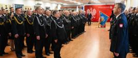 Uroczysta zbiórka w sali konferencyjnej. Po lewej stronie stoi zwarty szyk strażaków w czarnych mundurach galowych, sznurach galowych i garnizonowych czapkach. Po prawej stronie, tyłem do obiektywu, stoi strażak składający meldunek. W tle widoczna jest czerwona ścianka z godłem Polski i napisem „Państwowa Straż Pożarna”.