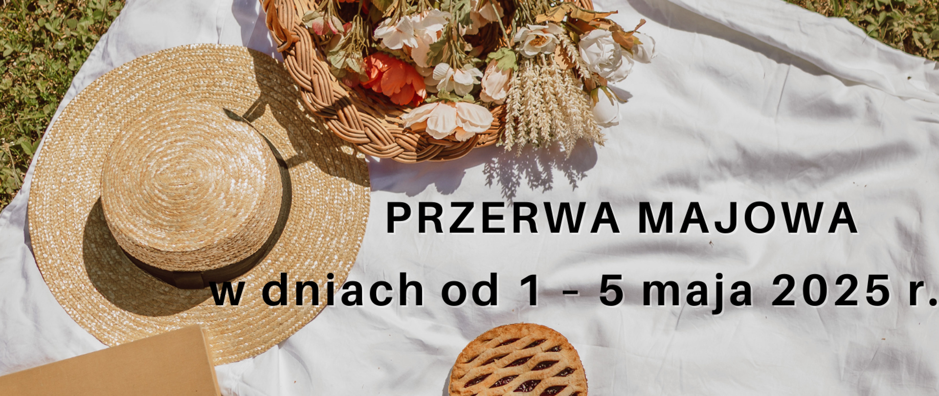 Na białym kocu piknikowym, rozłożonym prawdopodobnie na trawie, znajdują się różne przedmioty sugerujące majówkę. W lewym górnym rogu leży słomkowy kapelusz z ciemnym paskiem wokół ronda, rzucający cień na koc. Obok niego, po prawej stronie, stoi wiklinowy koszyk wypełniony różnorodnymi kwiatami w odcieniach bieli, różu i pomarańczy, a także suszonymi trawami. W prawym dolnym rogu widać okrągłe ciasto z kratką na wierzchu. Na środku obrazu, dużymi czarnymi literami, umieszczony jest tekst "PRZERWA MAJOWA", a pod nim mniejszymi literami "w dniach od 1 - 5 maja 2025 r.". Kompozycja sugeruje relaksującą atmosferę majowego wypoczynku na świeżym powietrzu.