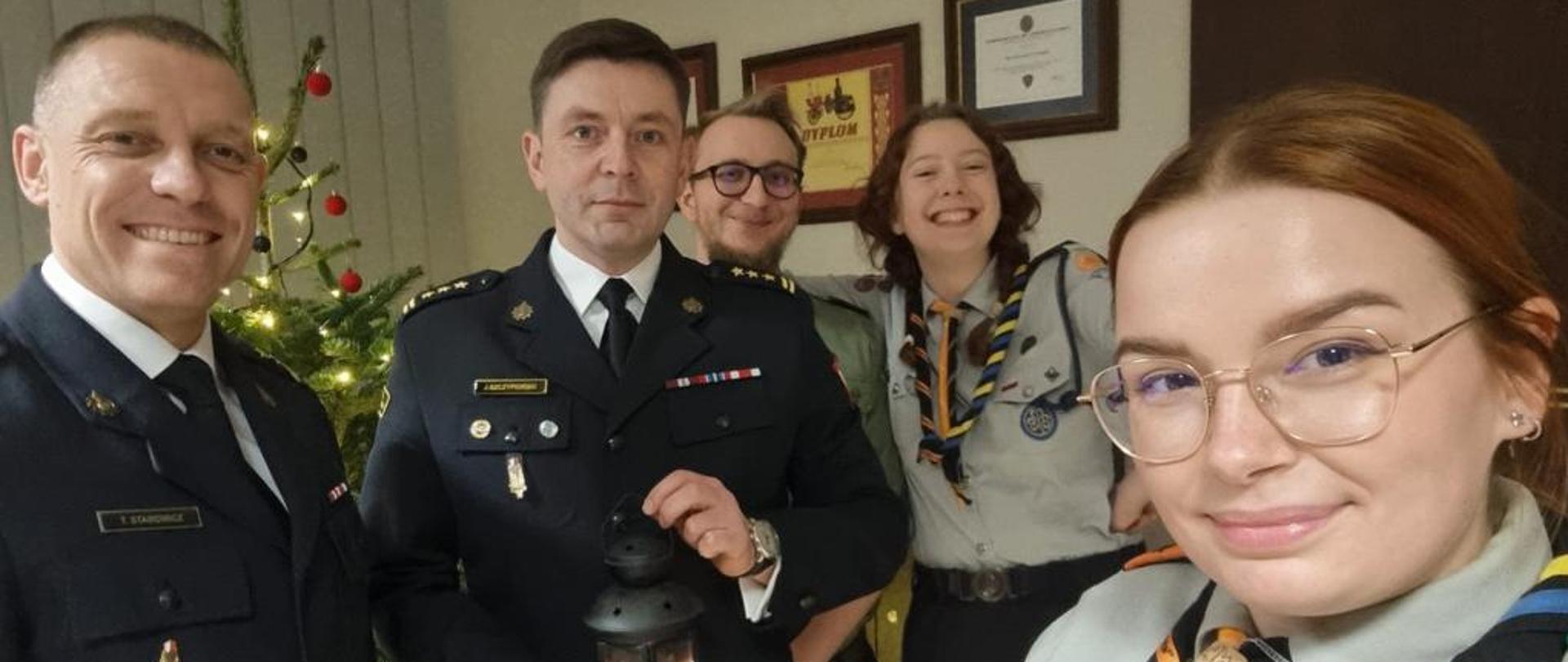 Komendant Miejski PSP st. bryg. Jacek Szczypiorski, Zastępca Komendanta Miejskiego PSP bryg. Tomasz Starowicz i harcerze Hufca ZHP Siemianowice Śląskie na wspólnym zdjęciu pamiątkowym po przekazaniu Betlejemskiego Światełka Pokoju. Komendant st. bryg. Jacek Szczypiorski trzyma w rękach otrzymane światełko pokoju.