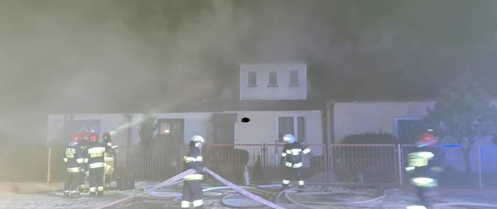 Na zdjęciu widać strażaków pracujących rozwijających linię gaśniczą przed silnie zadymionym budynkiem. Dym uniemożliwiał dokładne wykonanie zdjęcia. Zdjęcie wykonano w porze nocnej. Kilku strażaków stoi przed wejściem na posesję sprawdzając gdzie się dokładnie pali.