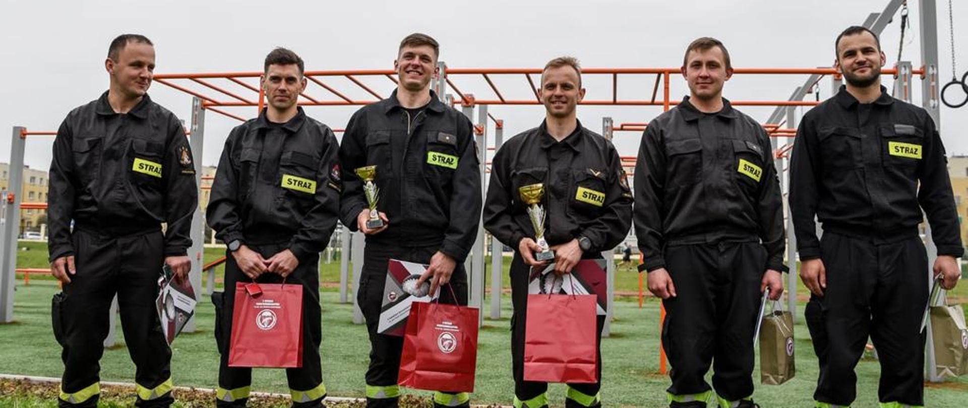 Sześciu strażaków stoi na boisku trawiastym, w rękach trzymają nagrody za udział w imprezie.