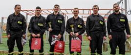 Sześciu strażaków stoi na boisku trawiastym, w rękach trzymają nagrody za udział.