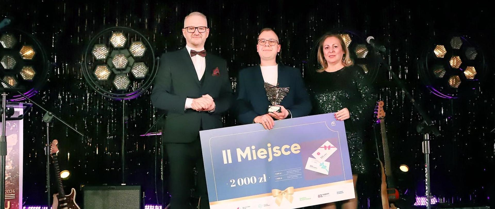Trzy osoby ubrane w eleganckie stroje, stoją na czerwonym dywanie na tle błyszczącej czarnej ściany. Stojący w środku mężczyzna trzyma voucher za zajęcie drugiego miejsca w kwocie 2000 zł oraz statuetkę. 