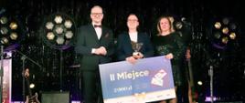 Trzy osoby ubrane w eleganckie stroje, stoją na czerwonym dywanie na tle błyszczącej czarnej ściany. Stojący w środku mężczyzna trzyma voucher za zajęcie drugiego miejsca w kwocie 2000 zł oraz statuetkę. 