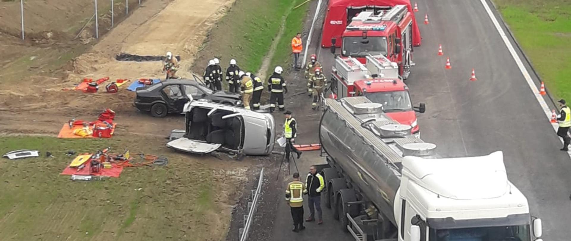 Widok na teren ćwiczeń z wiaduktu. Na pierwszym planie samochód ciężarowy cysterna i dwa uszkodzone auta osobowe, jedno leży na prawym boku, drugie stoi na kołach. W tle wozy strażackie stojące jeden za drugim wzdłuż drogi ekspresowej. 
