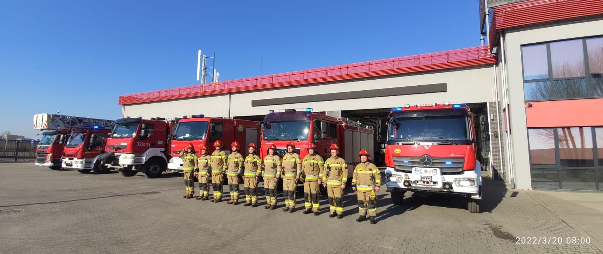 Zdjęcie wykonane przed jednostką ratowniczo – gaśniczą numer 2 w Radomiu. Na zdjęciu widać strażaków ustawionych w szeregu przed bramami garażowymi. Strażacy ubrani w ubrania specjalne w kolorze żółtym. Na głowach mają czerwone hełmy. Za nimi ustawione są pojazdy ratowniczo – gaśnicze w kolorze czerwonym. Pojazdy mają włączone alarmowe sygnały świetlne i dźwiękowe. Dowódca zmiany honory. Strażacy upamiętniają strażaków z Ukrainy którzy zginęli podczas działań wojennych. 