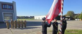 Strażacy na zbiórce do zdania służby, trzech strażaków ubranych w umundurowanie służbowe przygotowani do podniesienia flagi.