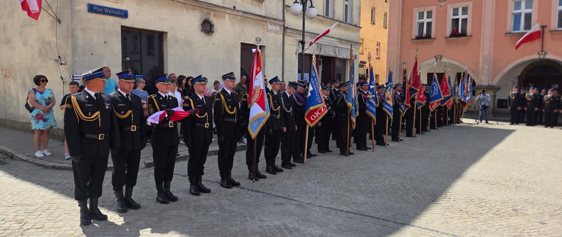 Strażacy w ciemnych mundurach galowych stojący w szyku na brukowanym rynku miejskim (Plac Wolności). Wzdłuż szeregu widoczne są liczne poczty sztandarowe ze sztandarami jednostek strażackich. Po lewej stronie, w tle, wydarzenie obserwuje grupa mieszkańców.