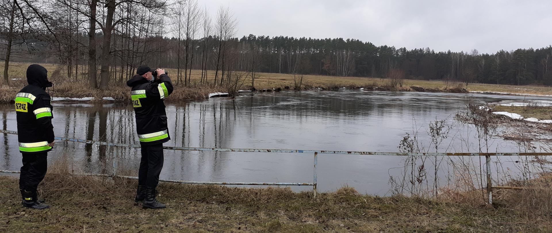 Na pierwszym planie widać rzekę oraz dwóch strażaków obserwujących stan wody. Jeden z nich patrzy przez lornetkę.