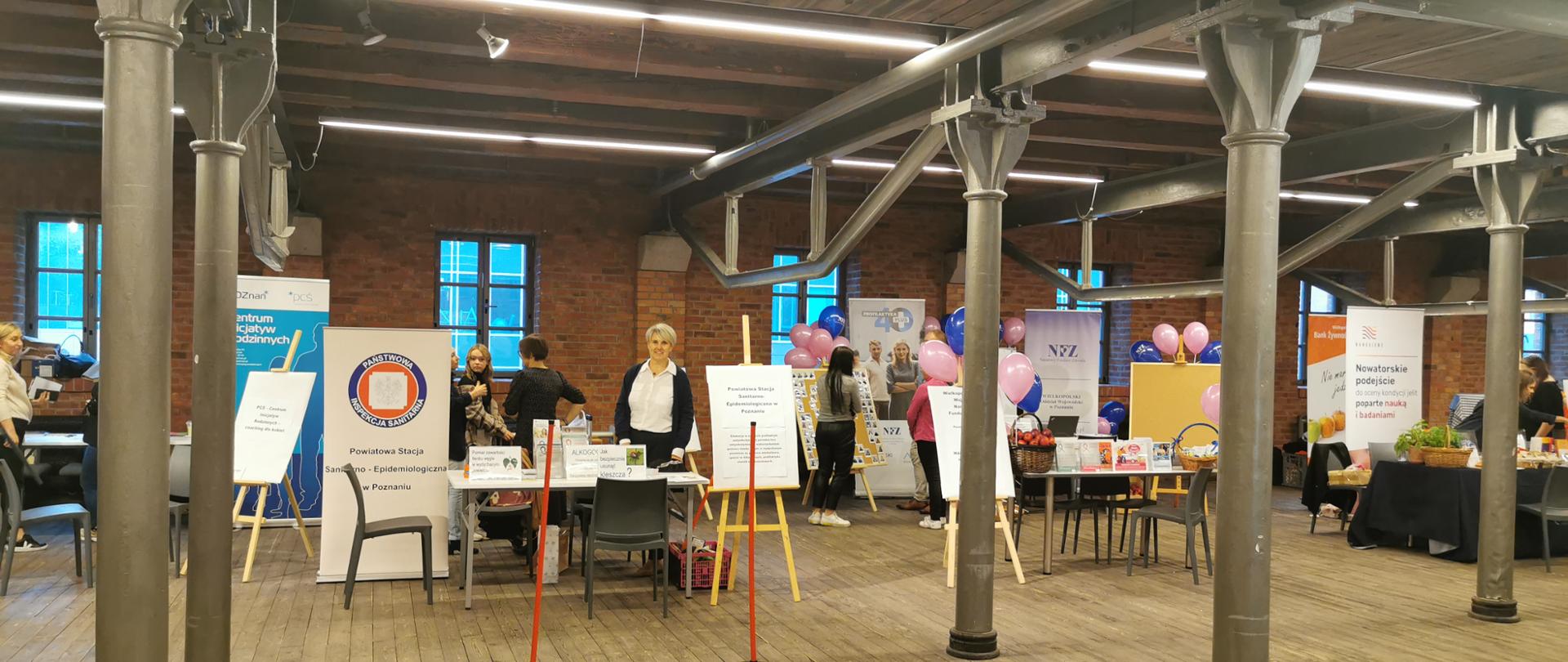 W dłużej Sali z kolumnami rozstawione stoiska instytucji i stowarzyszeń, na środku stoisko PSSE w Poznaniu.
