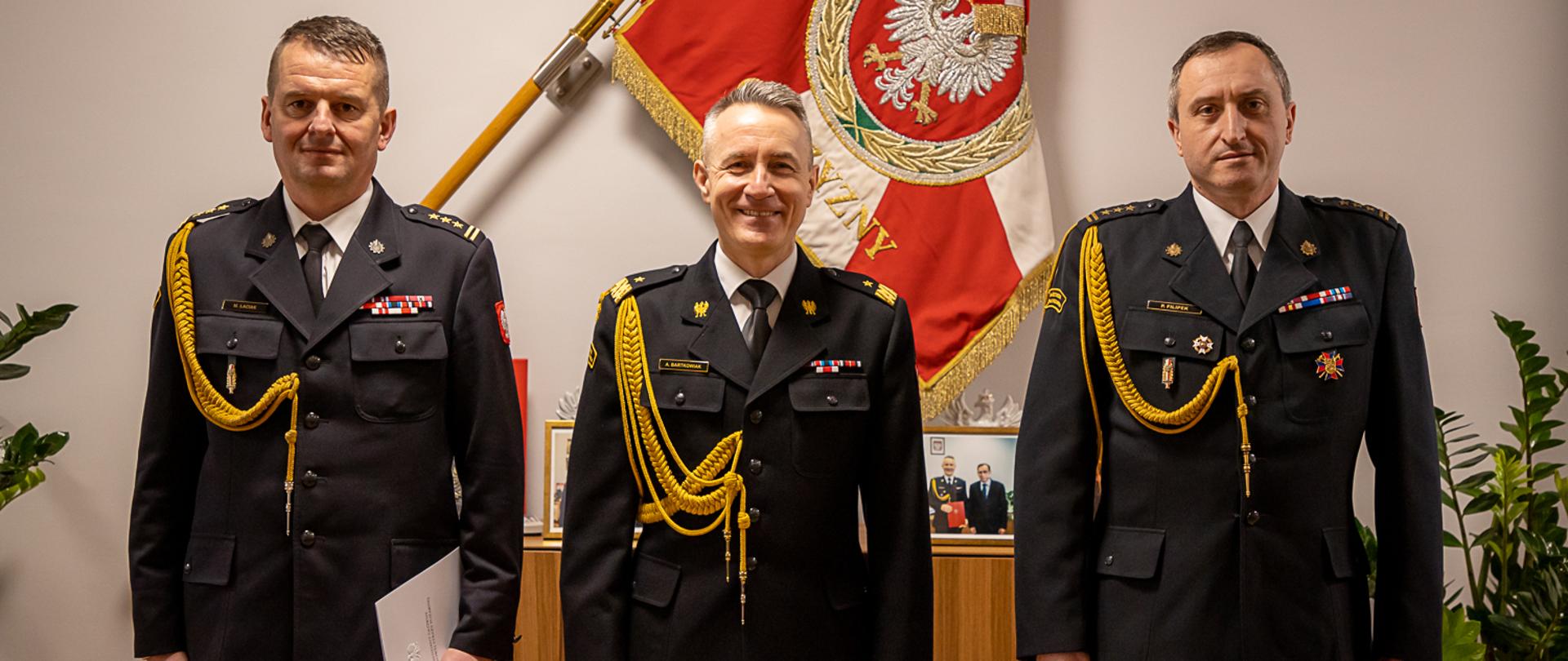 Na zdjęciu widać trzech strażaków. W środku stoi komendant główny PSP, w tle sztandar Komendy Głównej PSP.