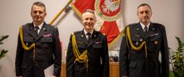 Na zdjęciu widać trzech strażaków. W środku stoi komendant główny PSP, w tle sztandar Komendy Głównej PSP.
