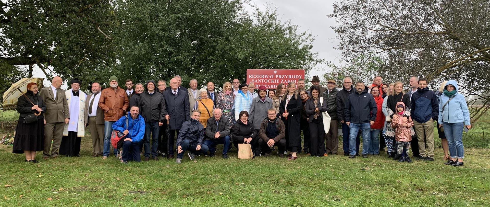 Grupa kilkudziesięciu osób pozuje do zdjęcia na tle tablicy z nazwą rezerwatu przyrody Santockie Zakole im. Ryszarda Popiela.