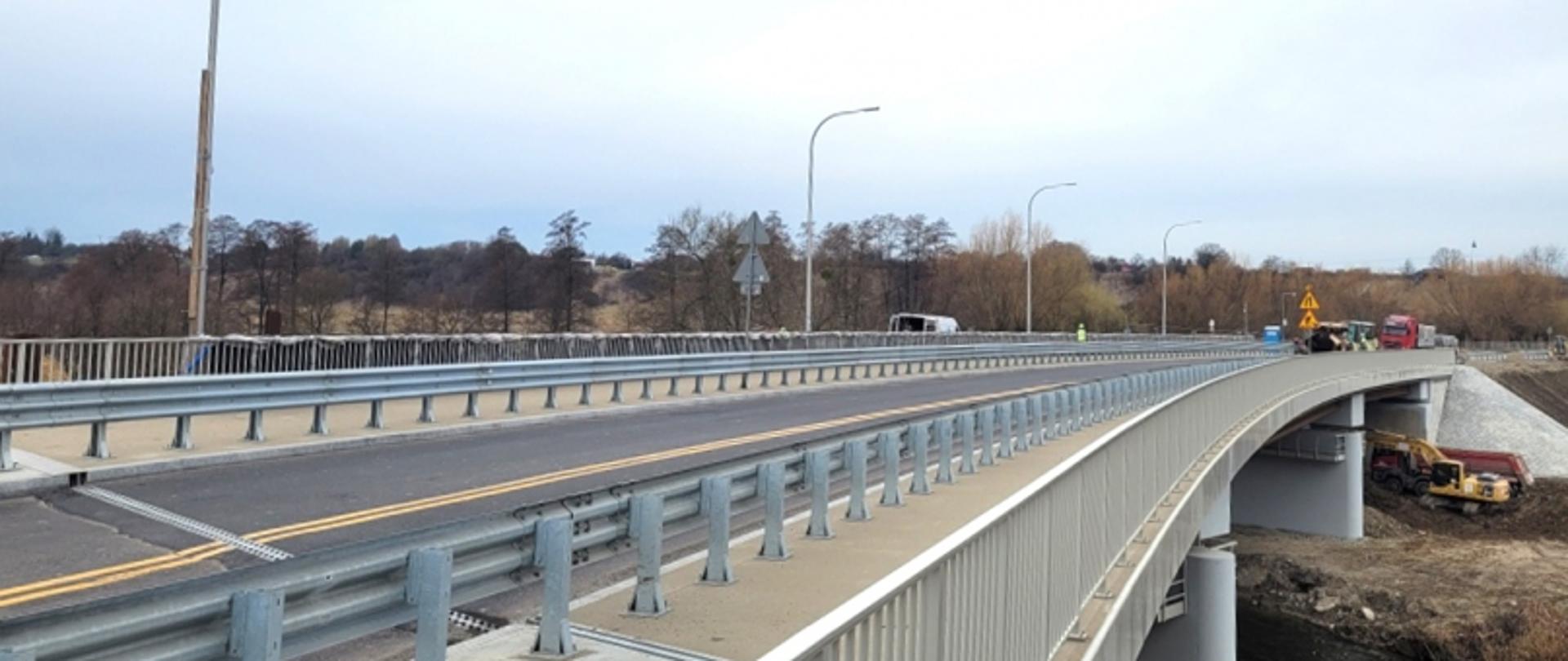 Nowy most na rzece Wieprz, który został dopuszczony do użytkowania przez nadzór budowlany. Konstrukcja mostu przebiega nad rzeką, po obu stronach zabezpieczona barierkami. W tle widoczne są pojazdy budowlane oraz oznakowanie drogowe, a po lewej stronie krajobraz z drzewami.
