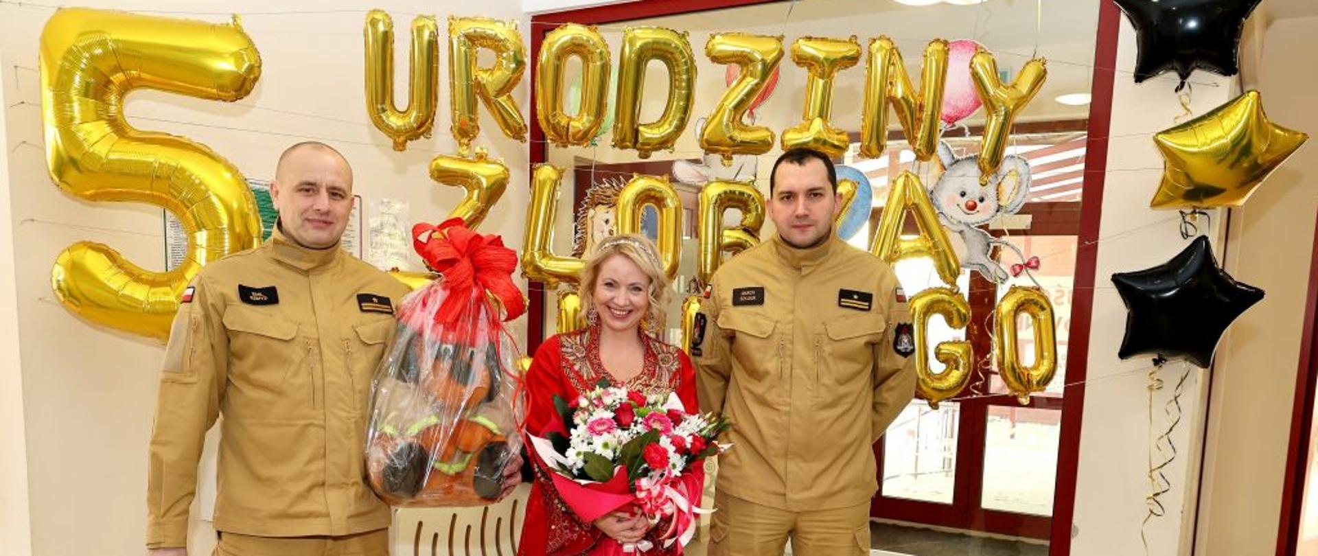 Na zdjęciu widać komendanta powiatowego psp w Ustrzykach Dolnych i jego zastępce w ubraniach koszarowych piaskowych, oraz kobietę z kwiatami w czerwonej sukni. Za nimi wisi napis z balonów "5 urodziny żłobka miejskiego"