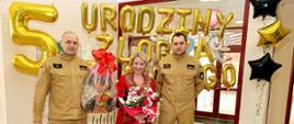 Na zdjęciu widać komendanta powiatowego psp w Ustrzykach Dolnych i jego zastępce w ubraniach koszarowych piaskowych, oraz kobietę z kwiatami w czerwonej sukni. Za nimi wisi napis z balonów "5 urodziny żłobka miejskiego"