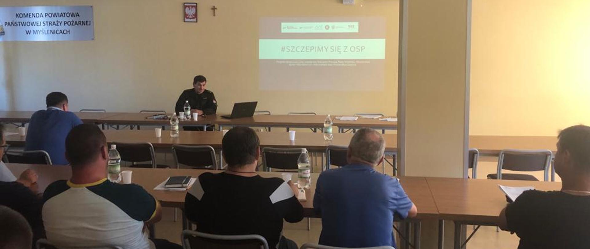 Zdjęcie przedstawia salę szkoleniową w Komendzie Powiatowej PSP w Myślenicach. Na ścianie z rzutnika wyświetlona jest nazwa programu. Z-ca Komendanta Powiatowego przedstawia projekt siedzącym druhom z jednostek OSP.
