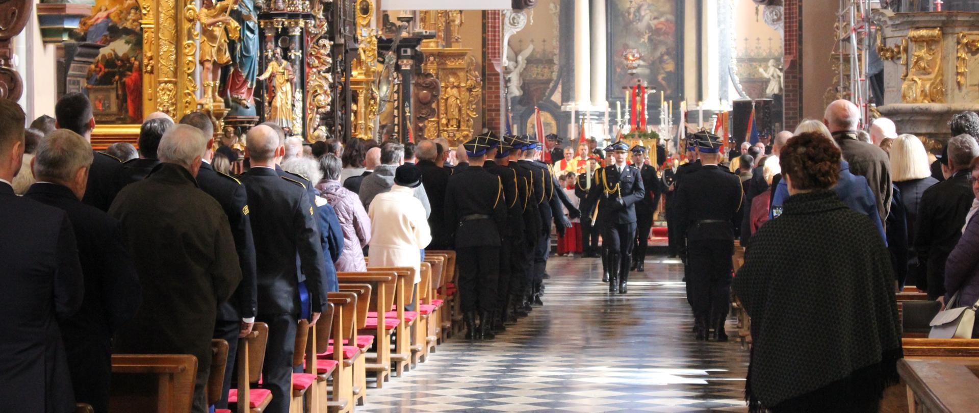 Wnętrze dużego kościoła, zdobione kolumny po prawej i lewej. ławeczki po prawej i lewej i ludzie w ławkach. W oddali ołtarz i strażacy w galowych mundurach.