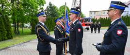 Na pierwszym planie trzech strażaków między dwoma dokonywany jest uścisk ręki dalej poczet sztandarowy oraz w tle inni strażacy i samochód pożarniczy