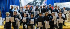Zdjęcie grupowe wykonane na scenie z drewnianą podłogą. Duża grupa dzieci i młodzieży w strojach galowych (głównie czarnych koszulach i spodniach) stoi w dwóch rzędach, trzymając w rękach dyplomy oraz białe i czarne torby upominkowe. Za nimi stoją dorośli mężczyźni, prawdopodobnie organizatorzy i jurorzy. W tle widoczna jest niebieska kurtyna, baner z logo konkursu przedstawiającym akordeon oraz roll-up z napisem "SCANDALLI". W dolnym rogu zdjęcia znajduje się logotyp "Twoja Słupca.pl".