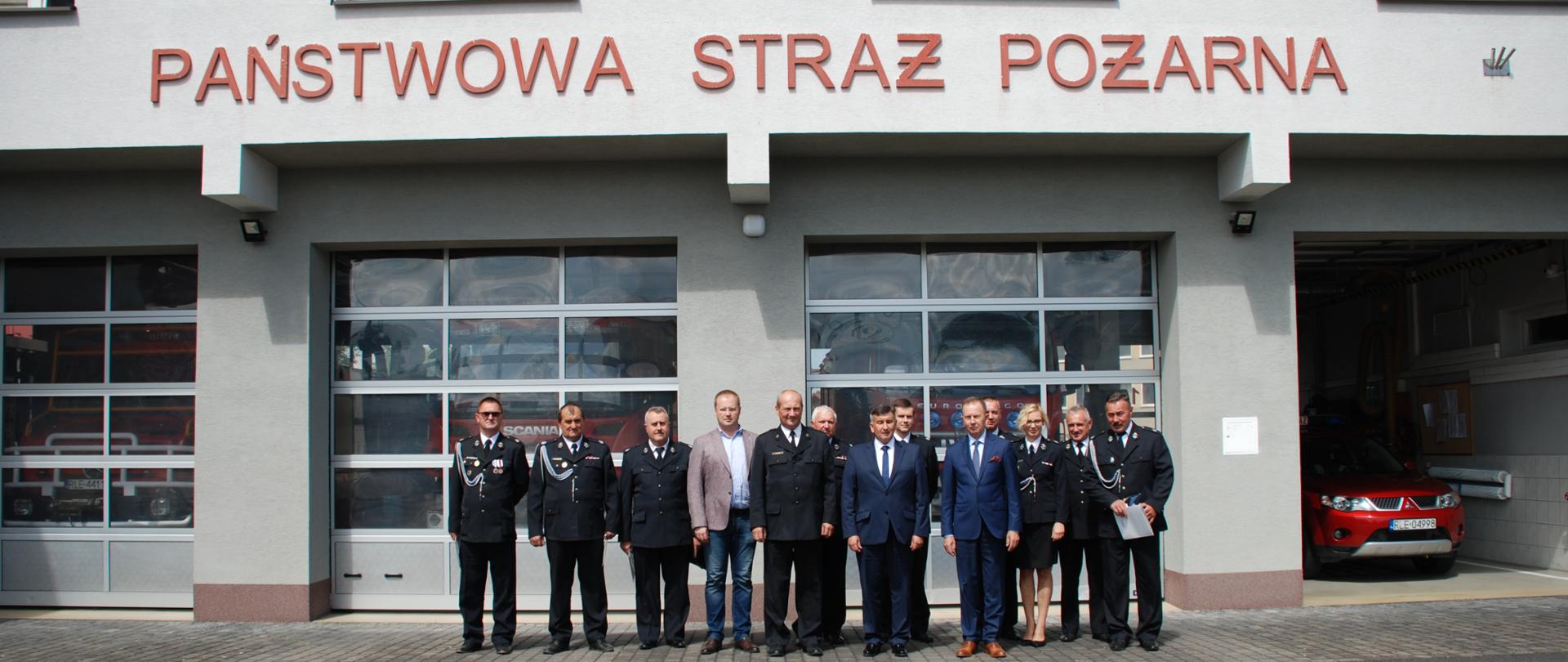Na zdjęciu widzimy druhów Ochotniczej Straży Pożarnej, komendanta Powiatowego Państwowej Straży Pożarnej w mundurach galowych oraz posła na Sejm RP Pana Jerzego Paula, zastępcę prezesa Wojewódzkiego Funduszu Ochrony Środowiska i Gospodarki Wodnej Pana Bogusława Kidę, radnego Sejmiku Województwa podkarpackiego Pana Karola Ożoga. Zebrani pozują do zdjęcia na tle budynku Komendy Powiatowej Państwowej Straży Pożarnej. 