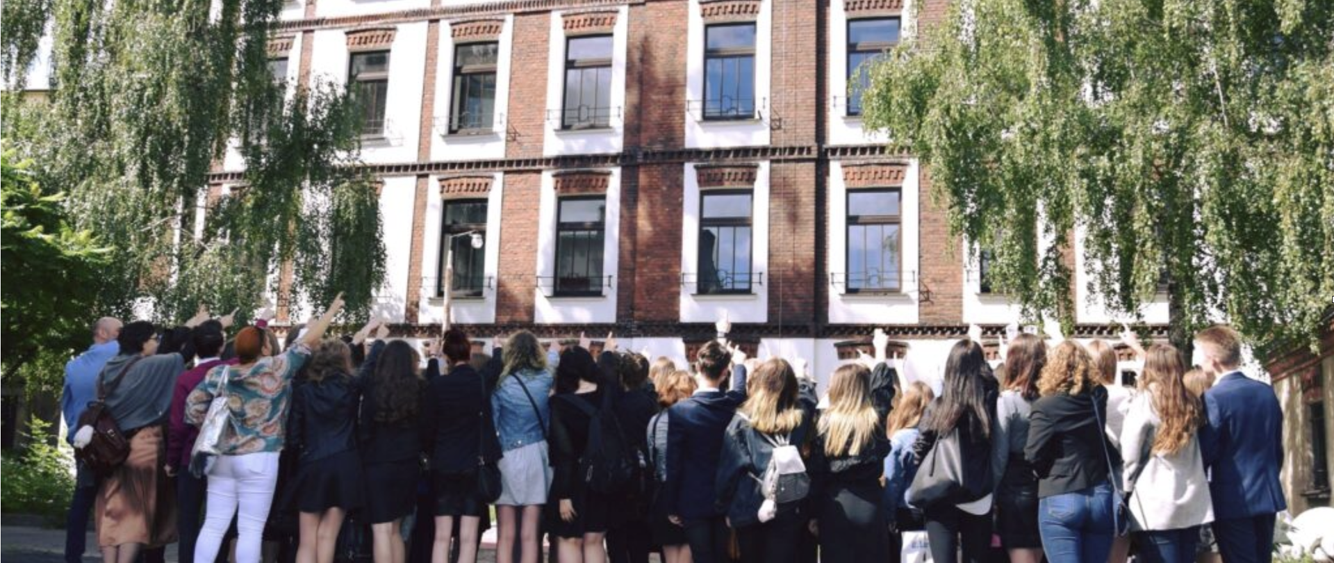 Przed frontem szkoły stoi tyłem do obserwatora grupa uczniów i nauczycieli, którzy wznoszą do góry ręce i wskazują na budynek