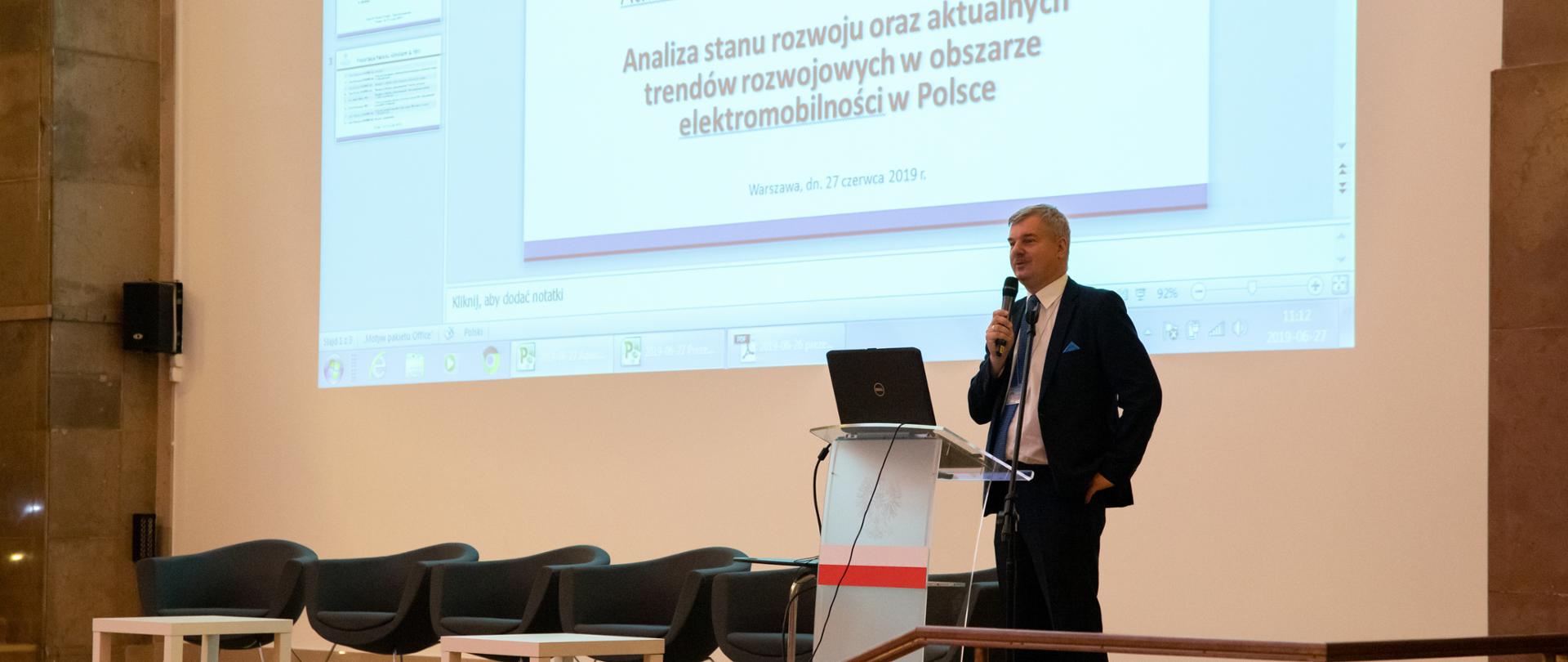 Prezentacja raportu o elektromobilności. Mężczyzna stoi na scenie. Za nim na akranie wyświetla się slajd tytułowy prezentacji.