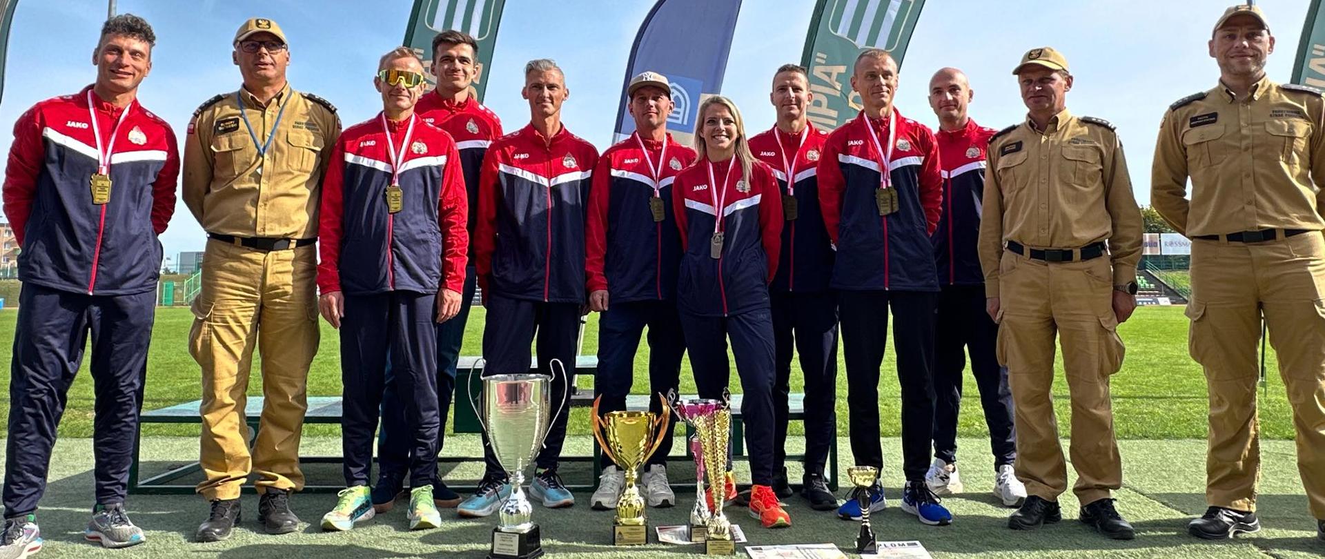 Grupowe zdjęcie drużyny PSP w czerwono-granatowych dresach z medalami oraz trofeami ustawionymi na murawie. Obok stoją funkcjonariusze w beżowych mundurach PSP, a w tle widoczne flagi i trybuny stadionu.