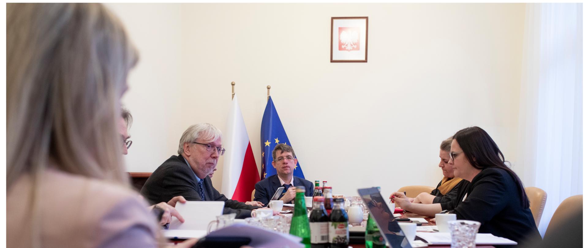 W pokoju przy stole siedzi grupa osób. Za nimi przy ścianie stoją dwie flagi - Polski i UE. Nad nimi wisi godło Polski.