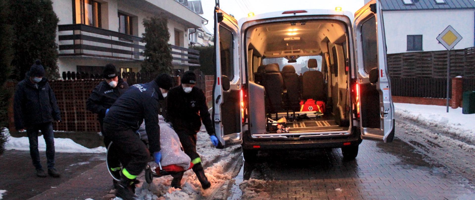 druhowie straży pożarnej pomagają w transporcie osoby niepełnosprawnej do punktu szczepień. Na drodze obok domu stoi specjalistyczny bus do przewozu osób poruszających się na wózku. Tylne drzwi samochodu są otwarte, a strażacy przenoszą seniorkę na wózku przez zaspę śniegu. Po bokach zdjęcia widać budynki mieszkalne. 