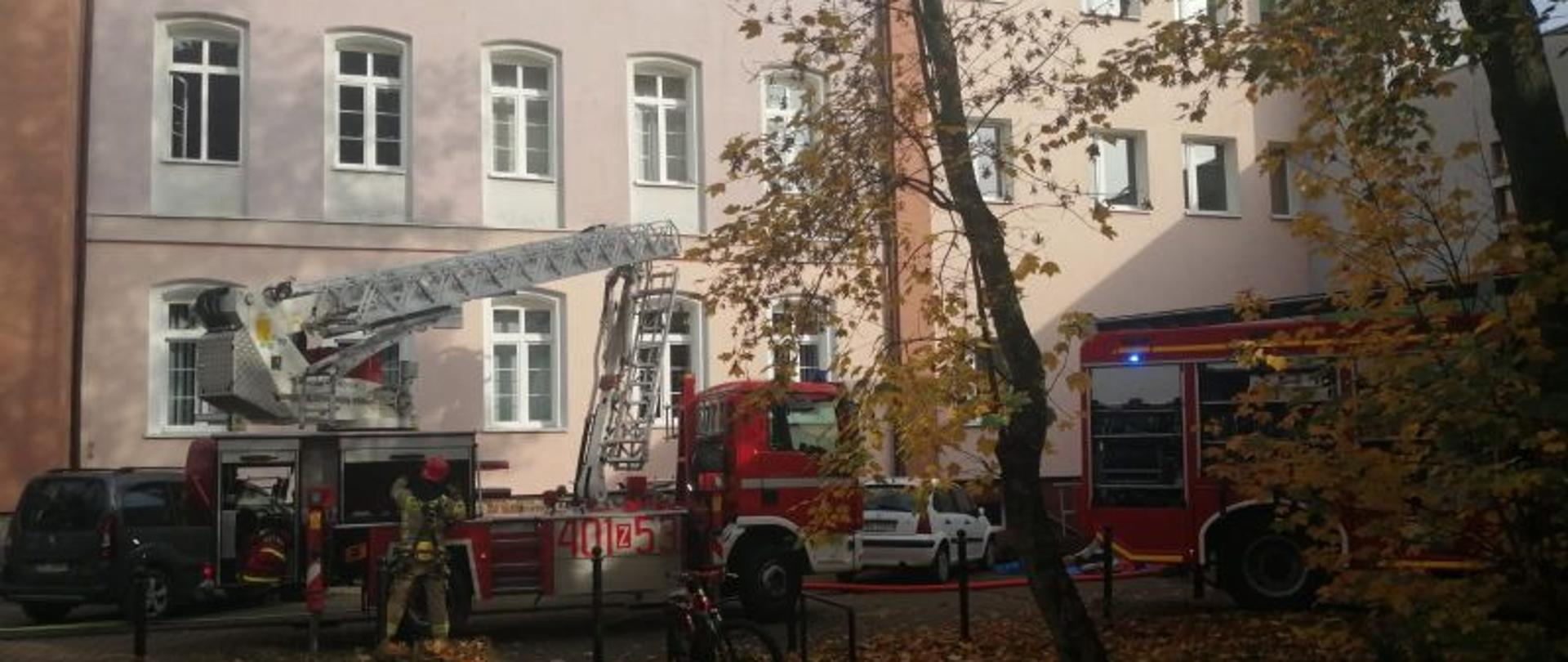 Widok na liceum z dalszej perspektywy. Widać podnośnik i stojącego przy nim strażaka. Widok przysłaniają drzewa. Budynek ma jasne kolory. Jest ładna jesienna pogoda.