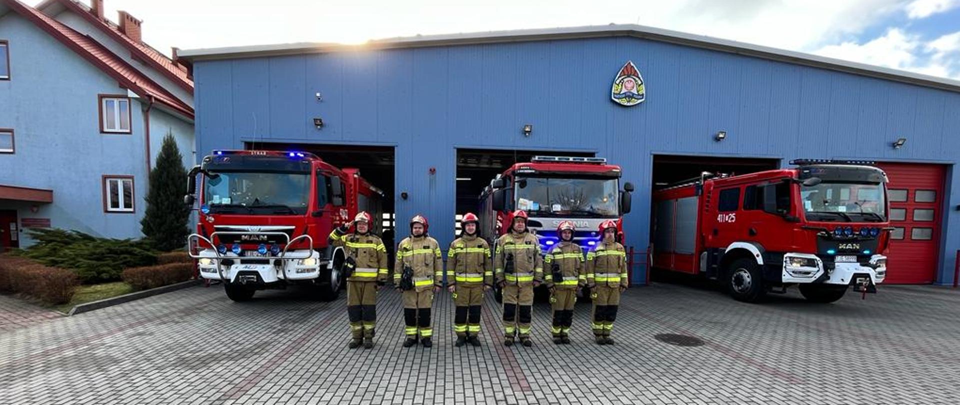 Zdjęcie obrazuje strażaków w umundurowaniu bojowym, którzy przed garażem jednostki ratowniczo-gaśniczej oddają hołd ofiarom katastrofy smoleńskiej. W otwartych bramach garażowych widać samochody ratowniczo-gaśnicze z włączonymi sygnałami świetlnymi. Jeden ze strażaków oddaje honor.