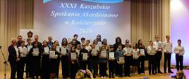 Na zdjęciu odświętnie ubrani uczniowie, którzy zostali laureatami XXXI Kaszubskich Spotkań Akordeonowych. Zdjęcie zostało wykonane podczas wręczenia nagród. Tło obrazka w odcieniach beżu. W tle na ścianie rzut niebieskiego slajdu z białymi napisem "XXXI Kaszubskie Spotkania Akordeonowe w Kościerzynie 2026". W oddali czarny fortepian, przed sceną zgromadzona publiczność.
