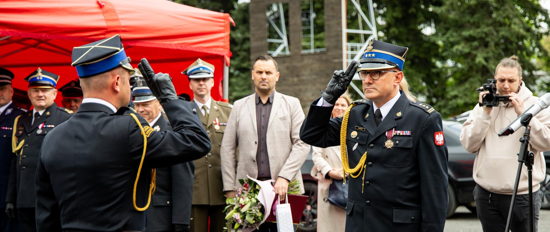 Zdjęcie przedstawia oficjalną, uroczystą ceremonię z udziałem służb mundurowych, odbywającą się na świeżym powietrzu. Głównym elementem kompozycji jest moment oddawania honorów przez dwóch wysokich rangą funkcjonariuszy Państwowej Straży Pożarnej.