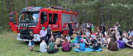 Na zdjęciu duże zgromadzenie dzieci i dorosłych na świeżym powietrzu, w większości siedzących na trawie i patrzących na duży, czerwony wóz strażacki zaparkowany po lewej stronie. Wóz strażacki ma napis „STRAŻ” z przodu i cyfry „352-21” po boku. Osoba w mundurze strażackim stoi w pobliżu wozu strażackiego, najwyraźniej prowadząc prezentację lub rozmowę z grupą. W tle widać las z licznymi drzewami.