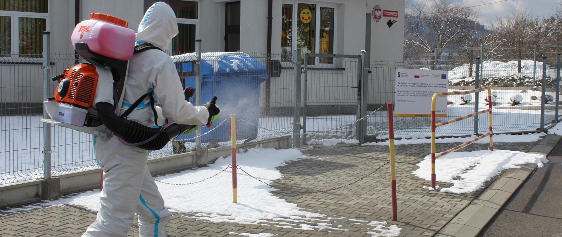 Strażak w specjalnym kombinezonie dezynfekuje okolicę szkoły opryskiwaczem spalinowym.