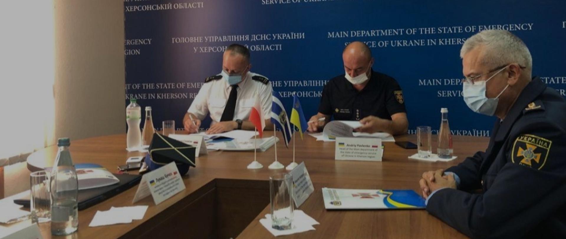 przy stole siedzą 3 osoby, 2 z nich podpisują dokumenty. Na stole znajdują się flagi Polski, Ukrainy i obwodu chersońskiego