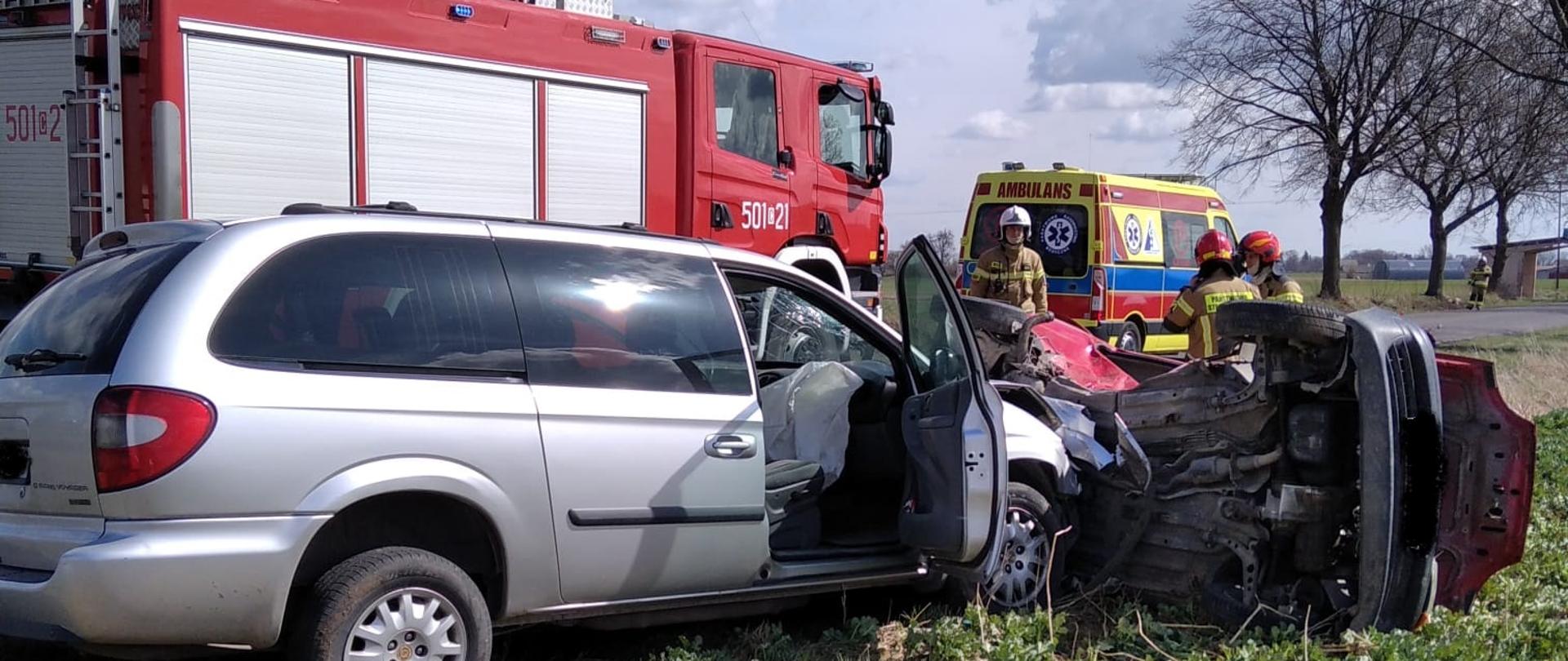 Zdjęcie przedstawia dwa samochody osobowe po wypadku koloru srebrnego oraz czerwonego. Na drugim planie pojazd pożarniczy koloru czerwnego oraz karetka pogotowia ratunkowego, oraz strażacy podczas działań ratowniczych