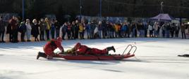 Zdjęcie przedstawia ćwiczących ratownictwo lodowe strażaków na lodowisku. W tle dzieci oglądające pokaz ratownictwa.