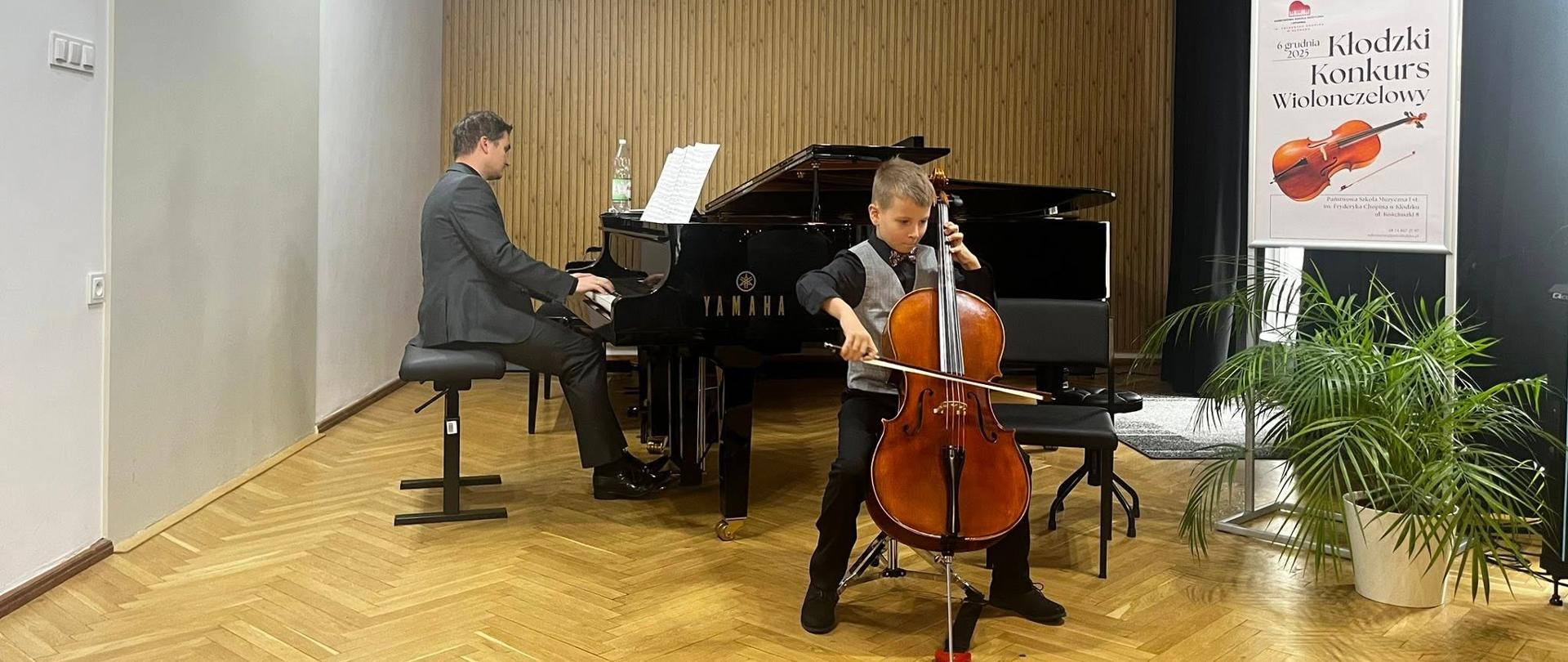 Wiolonczelista na scenie podczas wstępu, za nim przy fortepianie akompaniator.