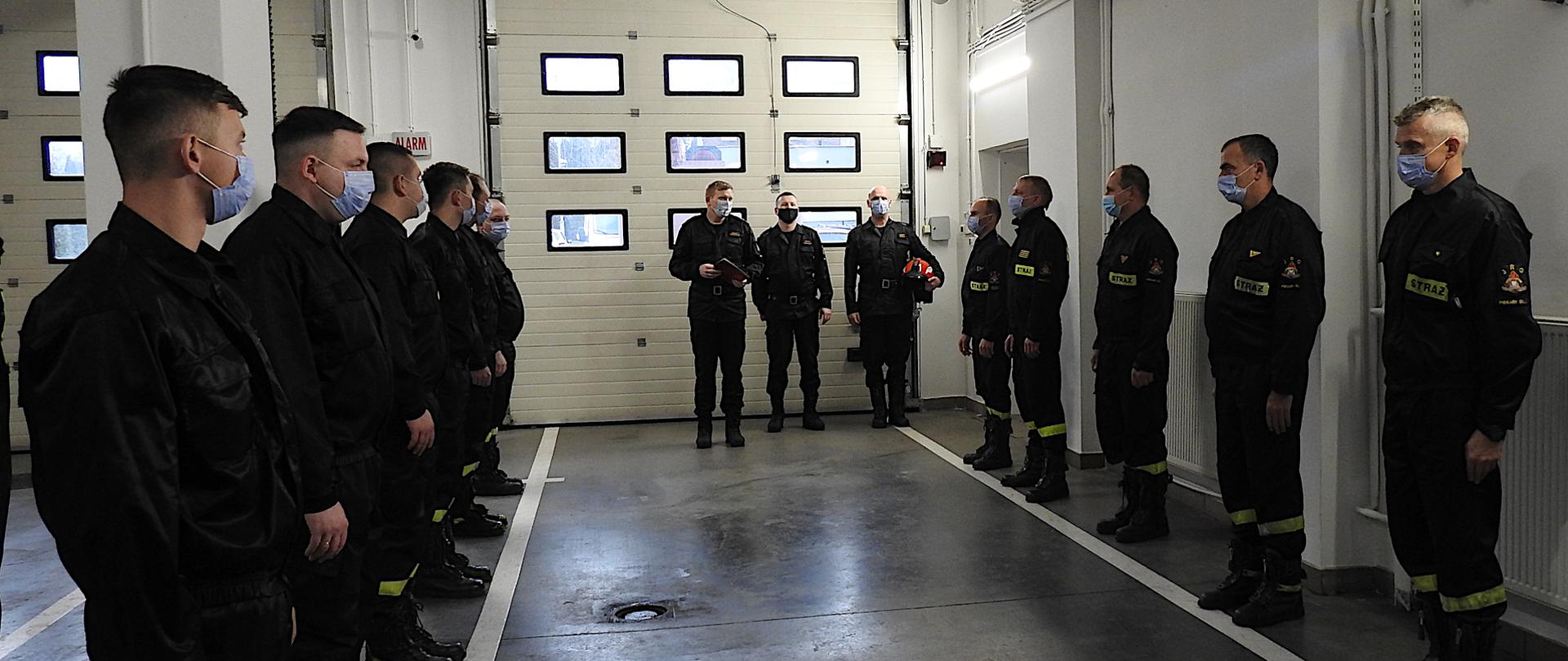 Zdjęcie przedstawia strażaków ustawionych w dwóch szeregach, stojący na przeciw siebie. Ubrani są w umundurowanie koszarowe i mają na twarzach maseczki chirurgiczne. Na końcu szpaleru stoją komendant miejski, jego zastępca oraz zastępca dowódcy jrg