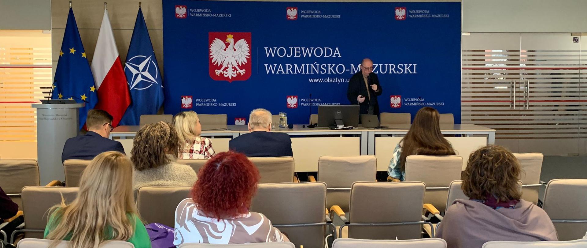 Zdjęcie przedstawia salę na której siedzi kilkanaście osób a w tle stoi mężczyzna i przemawia