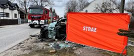 Samochód osobowy po dachowaniu przysłonięty parawanem z napisem straż, który zasłania ofiarę wypadku. Na drodze stoją samochody strażackie. Wokół rozbitego samochodu leżą potłuczone szyby i części karoserii.