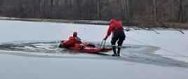 Na zdjęciu widoczni dwaj strażacy na skutym lodzie akwenie wodnym. Obaj w suchych skafandrach koloru pomarańczowego i czerwonego w czerwonych kaskach ochronnych. Jeden ratownik leży na saniach drugi asekurujący sanie oparty o podpory ewakuują z wody osobę poszkodowaną.
