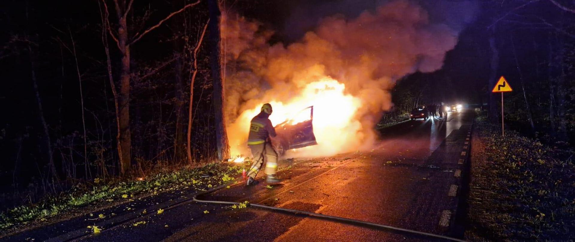 Na poboczu drogi pali się samochód osobowy. Widać kłęby dymu. Strażak za pomocą wody przystępuje do gaszenia pożaru.