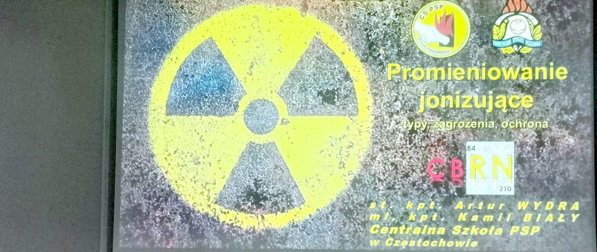 Po lewej stojący przy stole konferencyjnym funkcjonariusz CS PSP referujący wykład u góry widoczny wyświetlony na ekranie multimedialnym slajd prezentacji