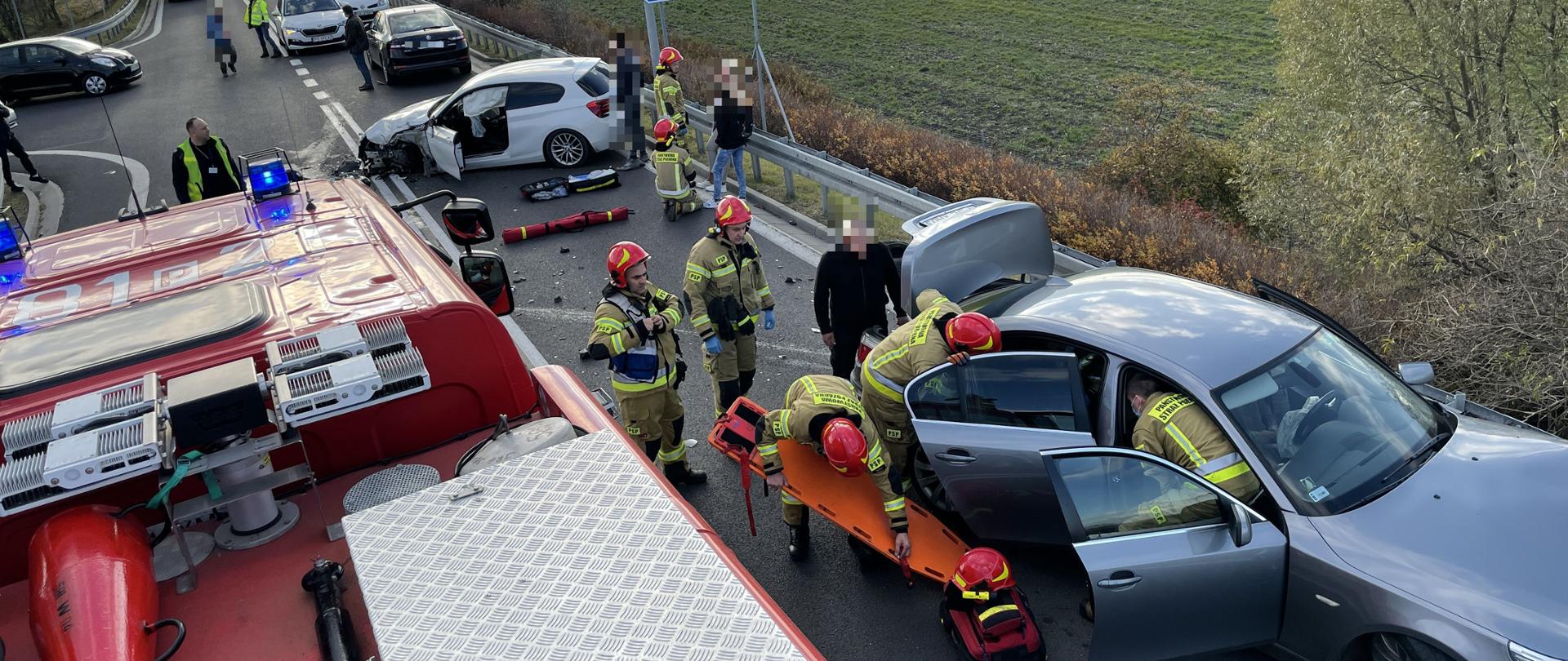 na zdjęciu widać uszkodzone dwa samochody jeden koloru białego drugi koloru szarego wokół samochodów znajdują się strażacy ze sprzętem medycznym obok samochodu siwego samochód strażacki czerwony jeden ze strażaków trzyma deskę ortopedyczna koloru pomarańczowego natomiast dwóch strażaków znajduje się w środku auta siwego udziela pomocy poszkodowanej osobie, dalej przy białym samochodzie jest strażak oraz dwie osoby 