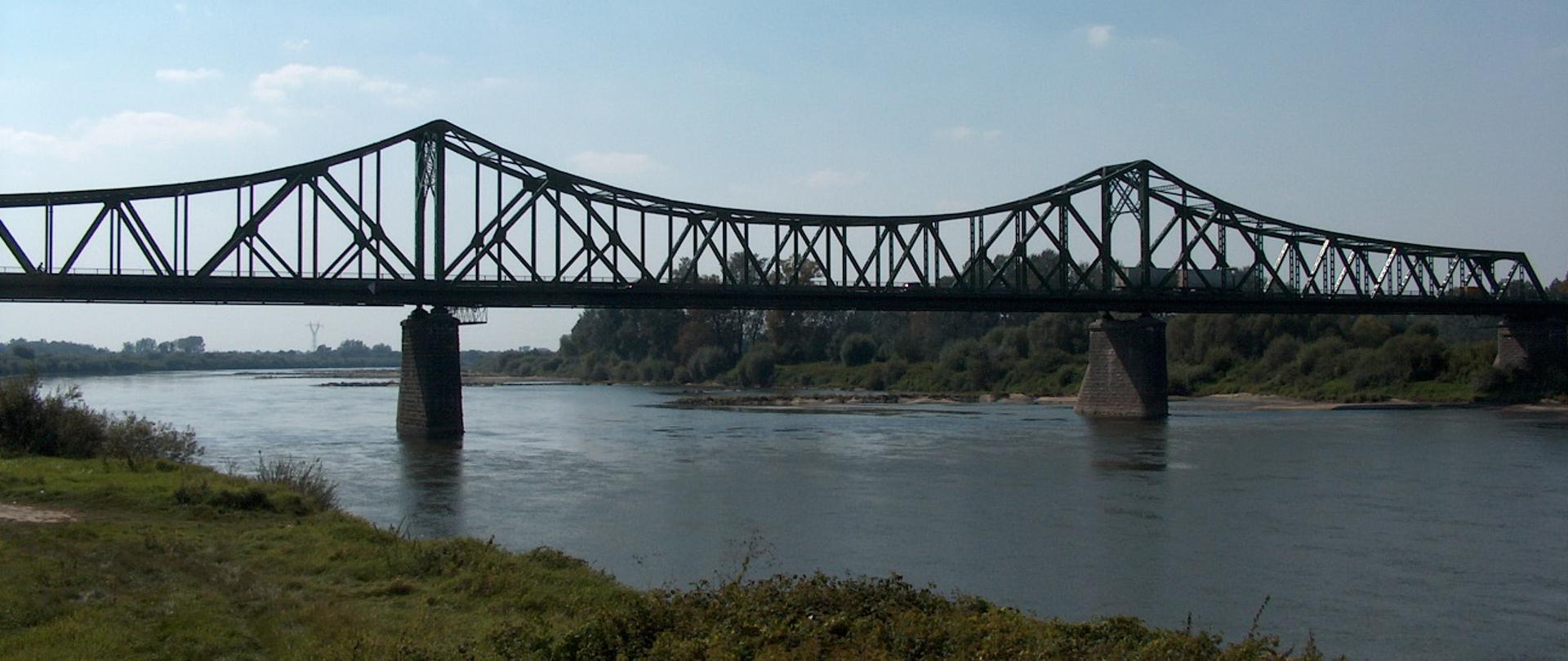 W środku rzeka Wisła, po obu stronach obrośnięte roślinami brzegi, most przez rzekę w Szczucinie o konstrukcji stalowej na kamiennych podporach 